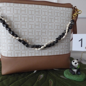 May include: A beige and brown crossbody bag with a repeating pattern. The bag features a decorative strap of black and white beads. A small panda figurine sits on a green leaf.