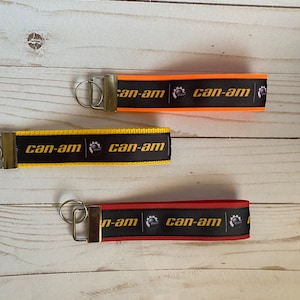 May include: Three keychains with black ribbon and gold metal rings. Each keychain has the text "can-am" in gold letters and a small black and gold logo. The keychains are in orange, yellow, and red.