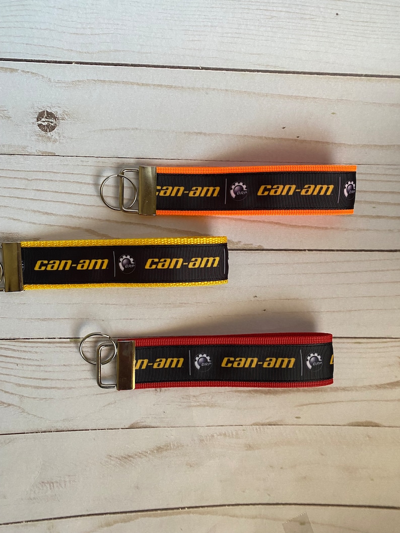 May include: Three keychains with black ribbon and gold metal rings. Each keychain has the text "can-am" in gold letters and a small black and gold logo. The keychains are in orange, yellow, and red.