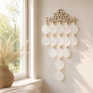 May include: A wall hanging featuring a laser-cut wooden top piece with an intricate scrollwork design. Suspended from this are multiple strands of translucent white circular discs interspersed with small, round, light-coloured wooden beads.