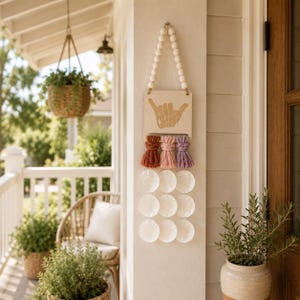 May include: A wall hanging featuring a square wooden plaque with a shaka hand sign graphic, suspended by a string of white wooden beads. Below the plaque are three colourful yarn tassels in rust, pink, and purple, with nine white shell discs.