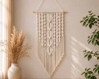 Puede incluir: Un tapiz de macramé de color crema con intrincados patrones de nudos, que incluyen formas de diamante centrales y columnas verticales trenzadas, suspendido de un listón de madera horizontal con un colgador triangular.