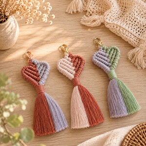 Puede incluir: Tres llaveros de macramé en forma de corazón con cierres de mosquetón dorados. Cada llavero presenta un diseño de cordón bicolor y una borla bicolor a juego. Las combinaciones de colores incluyen terracota y lavanda, crema y terracota, y lavanda y verde salvia.