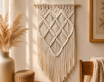 Puede incluir: Un tapiz de macramé color crema con un patrón de nudos en forma de diamante, suspendido de un palo de madera horizontal con un cordón a juego. La sección inferior consiste en flecos largos, rectos y sin nudos.