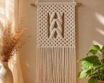 Puede incluir: Un tapiz de macramé de color crema suspendido de un listón de madera horizontal con un cordón blanco. La pieza presenta un patrón trenzado central rodeado por una cuadrícula de nudos, rematado con largos flecos rectos que cuelgan abajo.