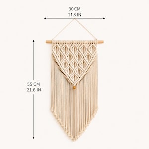 Puede incluir: Un tapiz de macram&eacute; color crema con un patr&oacute;n de nudos en forma de diamante, suspendido de un palo de madera con una cuenta de madera en la punta inferior. La pieza mide 30 cm de ancho y 55 cm de largo total.