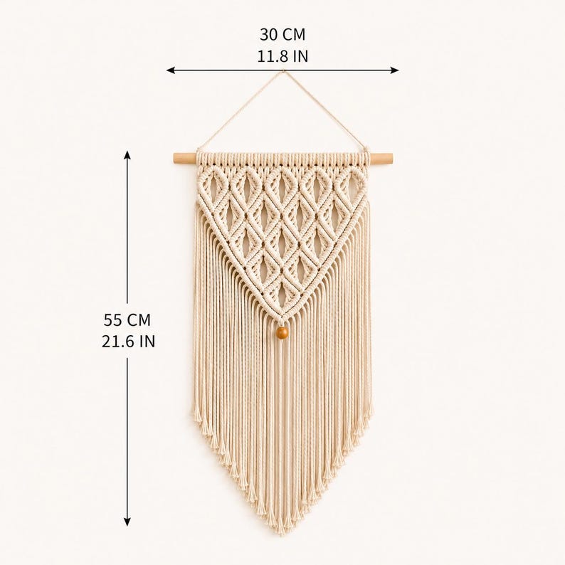 Puede incluir: Un tapiz de macram&eacute; color crema con un patr&oacute;n de nudos en forma de diamante, suspendido de un palo de madera con una cuenta de madera en la punta inferior. La pieza mide 30 cm de ancho y 55 cm de largo total.