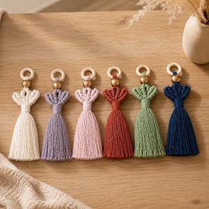 May include: Six macrame tassels in cream, lavender, light pink, terracotta, sage green, and navy blue. Each tassel features a wooden ring at the top, a metallic gold bead, a decorative knotted fan pattern, and a long, straight fringe base.