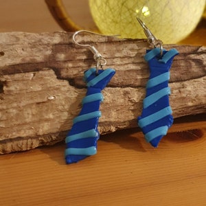 Puede incluir: Un par de pendientes hechos a mano con forma de corbata. Los pendientes son principalmente azules con rayas y detalles en azul claro. Cuelgan de ganchos plateados. Los pendientes se exhiben sobre un trozo de madera.