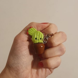 Op de afbeelding: Een groene cactus sleutelhanger met een lachend gezicht en rode bloemaccenten, in een bruine pot. De sleutelhanger is bevestigd aan een zilveren metalen sluiting. Een leuk, grillig accessoire.