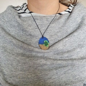 Puede incluir: Collar con colgante ovalado que presenta una escena oceánica pintada con cielo azul, arena color arena y una tortuga verde. El colgante está suspendido de un cordón negro. El collar se lleva sobre un suéter gris.