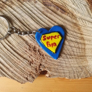 May include: A blue heart-shaped keychain with the words "Super Papa" in red letters inside a yellow diamond. The keychain is attached to a silver metal ring and chain.