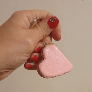 Könnte beinhalten: Ein rosa herzförmiger Schlüsselanhänger mit einem silberfarbenen Metallring. Der Schlüsselanhänger wird in einer Hand mit rotem Nagellack gehalten.
