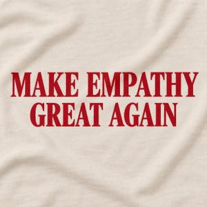 May include: A light beige fabric featuring the text "MAKE EMPATHY GREAT AGAIN" printed in a bold, dark red serif font. The material displays a soft, slightly wrinkled texture, with the lettering centred clearly across the surface of the textile.