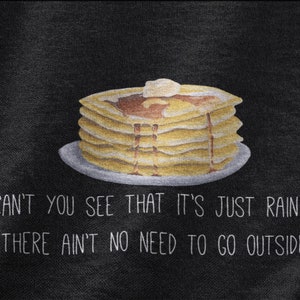 May include: A black fabric surface featuring a graphic illustration of a stack of golden-brown pancakes on a plate, topped with a pat of butter and syrup. Below the pancakes, white text reads: CAN'T YOU SEE THAT IT'S JUST RAININ' THERE AIN'T NO NEED TO GO OUTSIDE.