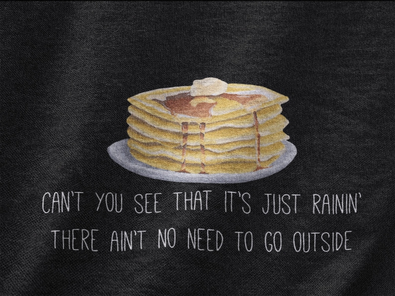 May include: A black fabric surface featuring a graphic illustration of a stack of golden-brown pancakes on a plate, topped with a pat of butter and syrup. Below the pancakes, white text reads: CAN'T YOU SEE THAT IT'S JUST RAININ' THERE AIN'T NO NEED TO GO OUTSIDE.