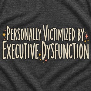 May include: A dark charcoal gray t-shirt featuring the cream-colored text "PERSONALLY VICTIMIZED BY EXECUTIVE DYSFUNCTION" in a stylized font. The design is accented with small, scattered four-pointed stars in shades of orange and pink.