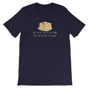 May include: A dark navy blue short-sleeve t-shirt featuring a graphic of a stack of pancakes with syrup and butter on a plate. Below the illustration, the text reads: CAN'T YOU SEE THAT IT'S JUST RAININ' THERE AIN'T NO NEED TO GO OUTSIDE.