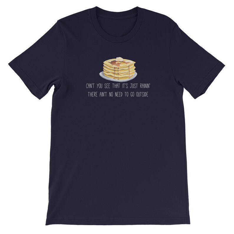 May include: A dark navy blue short-sleeve t-shirt featuring a graphic of a stack of pancakes with syrup and butter on a plate. Below the illustration, the text reads: CAN'T YOU SEE THAT IT'S JUST RAININ' THERE AIN'T NO NEED TO GO OUTSIDE.