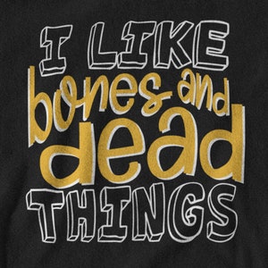 May include: A black fabric surface featuring the text I LIKE BONES AND DEAD THINGS. The words I LIKE and THINGS are written in a white outlined font, while the words bones and dead are printed in a solid gold color with a white drop shadow.