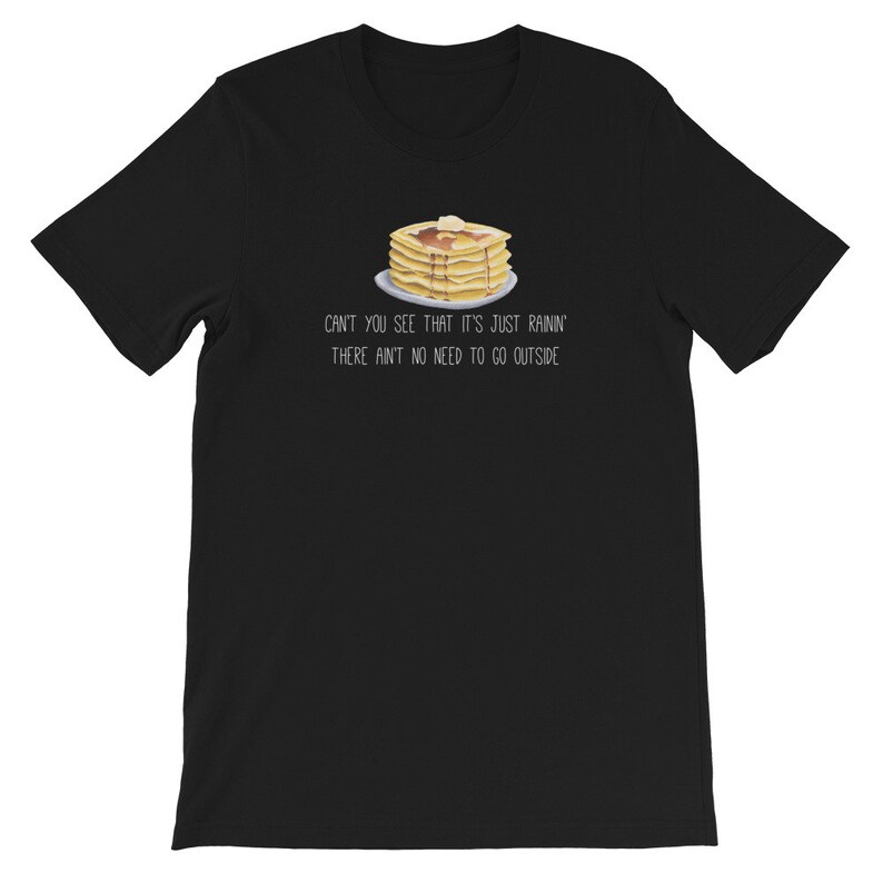 May include: A black short-sleeved t-shirt featuring a graphic of a stack of pancakes with syrup and butter on a plate. Below the illustration, the text reads: CAN'T YOU SEE THAT IT'S JUST RAININ' THERE AIN'T NO NEED TO GO OUTSIDE in white lettering.