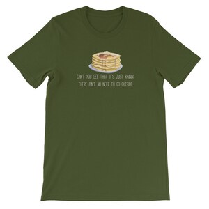 May include: An olive green short-sleeve t-shirt featuring a graphic of a stack of pancakes with syrup and butter on a plate. Below the illustration, the text reads: CAN'T YOU SEE THAT IT'S JUST RAININ' THERE AIN'T NO NEED TO GO OUTSIDE.