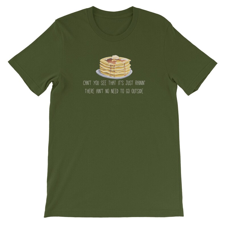 May include: An olive green short-sleeve t-shirt featuring a graphic of a stack of pancakes with syrup and butter on a plate. Below the illustration, the text reads: CAN'T YOU SEE THAT IT'S JUST RAININ' THERE AIN'T NO NEED TO GO OUTSIDE.