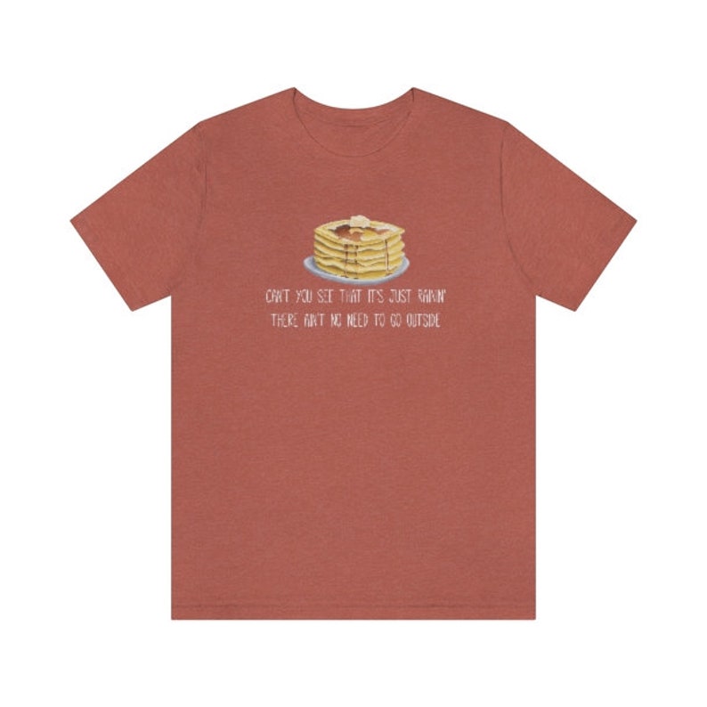 May include: A rust-colored short-sleeve t-shirt featuring a graphic of a stack of pancakes with syrup and butter on a plate. Below the graphic, the text reads: CAN'T YOU SEE THAT IT'S JUST RAININ' THERE AIN'T NO NEED TO GO OUTSIDE in white lettering.