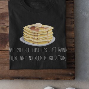 May include: A black t-shirt featuring a graphic of a stack of pancakes with syrup and a pat of butter on a plate. Below the illustration, the white text reads: CAN'T YOU SEE THAT IT'S JUST RAINING, THERE AIN'T NO NEED TO GO OUTSIDE.