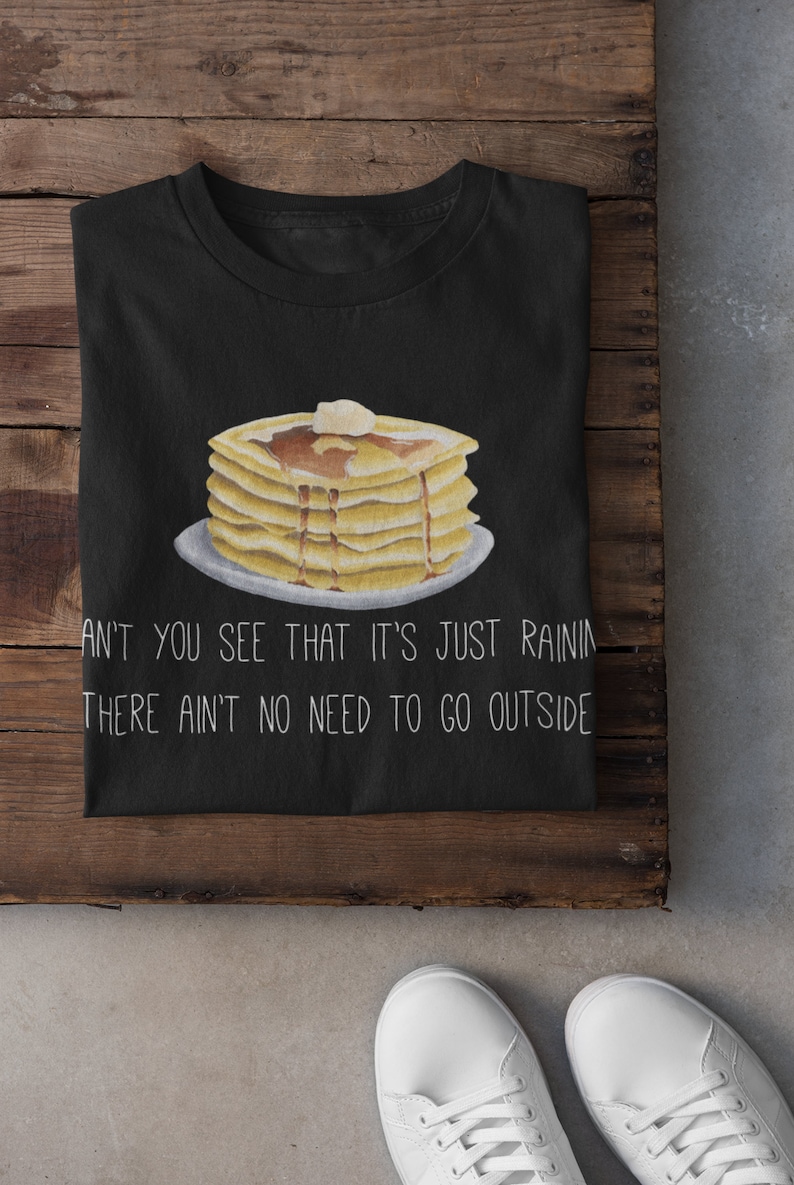 May include: A black t-shirt featuring a graphic of a stack of pancakes with syrup and a pat of butter on a plate. Below the illustration, the white text reads: CAN'T YOU SEE THAT IT'S JUST RAINING, THERE AIN'T NO NEED TO GO OUTSIDE.