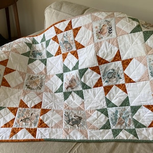 May include: A quilted blanket featuring a star pattern with alternating white, burnt orange, and sage green fabric blocks. The quilt includes square panels printed with illustrations of rabbits, ducks, and small birds surrounded by floral foliage.