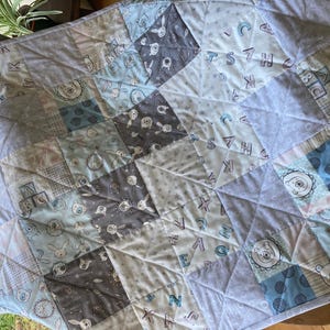May include: A patchwork quilt featuring a variety of square fabric panels in shades of blue, grey, and white. The patterns include illustrations of rabbits, bears, and alphabet blocks with the letters ABC, all stitched together with a diamond pattern.