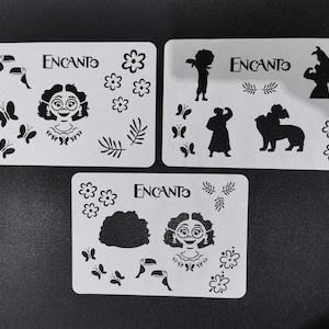 May include: Three white stencils with black cutouts featuring characters and objects from the Disney movie Encanto. The stencils include Mirabel, Bruno, a toucan, flowers, and leaves.