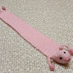 May include: A pink crocheted pig bookmark with black eyes. The bookmark is lying on a white textured surface.