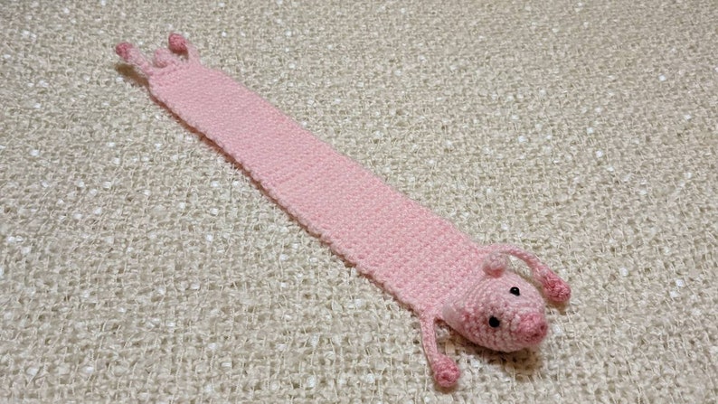 May include: A pink crocheted pig bookmark with black eyes. The bookmark is lying on a white textured surface.