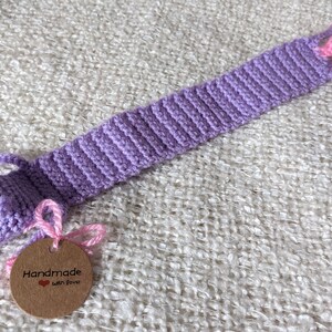 May include: Purple crocheted bookmark shaped like a pig with pink ears and nose. The bookmark has a brown tag that says "Handmade with love".