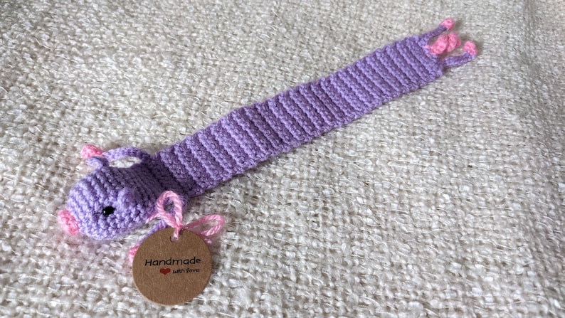 May include: Purple crocheted bookmark shaped like a pig with pink ears and nose. The bookmark has a brown tag that says "Handmade with love".