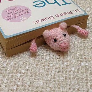 Pink crocheted pig bookmark peeking out from between the pages of a book.