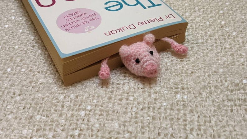 Pink crocheted pig bookmark peeking out from between the pages of a book.