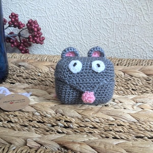 Puede incluir: Un ratón gris tejido a mano con ojos blancos, orejas rosas y una lengua rosa. El ratón está sobre una superficie tejida. Una botella de vidrio azul y bayas rojas están en el fondo. Pequeñas etiquetas están en primer plano.