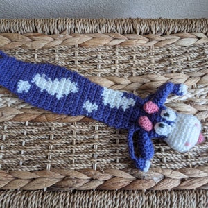 May include: A crocheted purple and white cow bookmark with pink ears and black eyes. The bookmark is lying on a woven brown surface.
