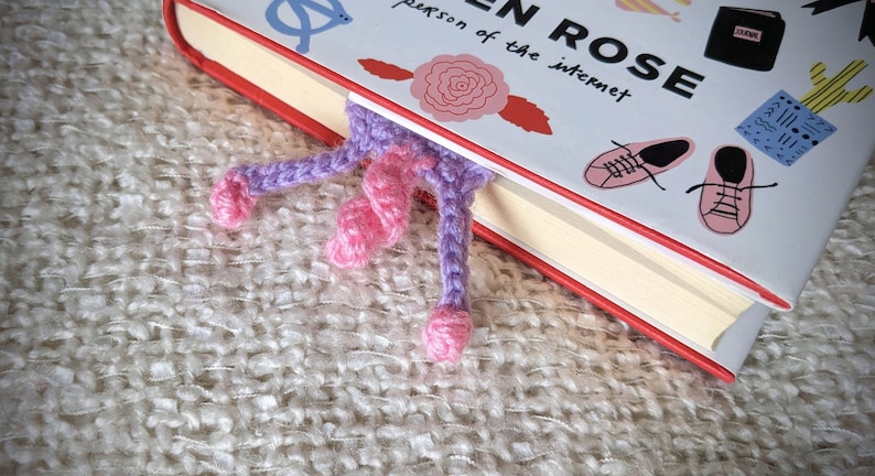 May include: A purple and pink crocheted bookmark with a curled tail, sticking out from the edge of a book titled "Modern Rose: The Person of the Internet".