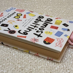 May include: A white book with a pink pig bookmark sticking out of it. The book cover has a colorful design with the title "Almost Everything" and the author's name "ARDEN ROSE".