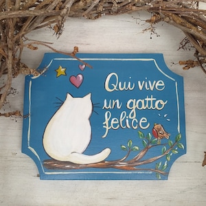 Può includere: Un cartello in legno dipinto a mano con sfondo blu raffigurante un gatto bianco, un uccello e la scritta italiana "Qui vive un gatto felice" (Qui vive un gatto felice). Il cartello è decorato con una stella e un cuore.