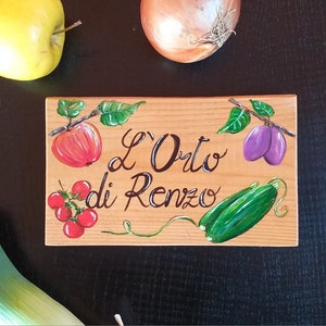 May include: A wooden sign with the text "L'Orto di Renzo" and painted illustrations of fruits and vegetables, including an apple, tomatoes, plums, and cucumbers. A yellow apple, an onion, and a leek are also visible.