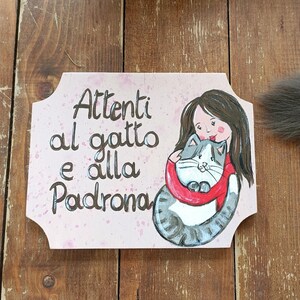 Könnte beinhalten: Ein rechteckiges Holzschild mit geschwungenen Kanten und einer handgemalten Illustration einer Person mit langem braunem Haar in einem roten Pullover, die eine grau-weiße Katze hält. Der Text lautet: Attenti al gatto e alla Padrona.