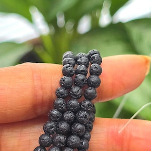 May include: Close-up of a black lava stone bracelet. The bracelet is made of small, round, porous beads strung together. The beads are a dark charcoal color, and the bracelet is held between two fingers against a blurred green background.