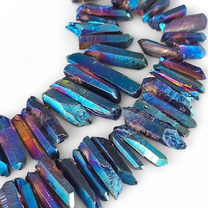 May include: Close-up of a collection of iridescent blue quartz crystal points. The crystals have a metallic sheen with hues of purple, gold, and teal. The crystals are arranged on a white surface.