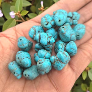 May include: A collection of turquoise-colored, irregular-shaped beads with dark brown veining. Each bead has a small hole, suggesting they are for stringing. The beads are held in a hand, with green foliage in the background.