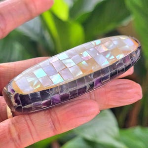 May include: An oblong, decorative object with a mosaic design. The top features a pattern of iridescent squares, transitioning from white to yellow. The sides are a mosaic of purple and gray squares. The object is held in a hand.